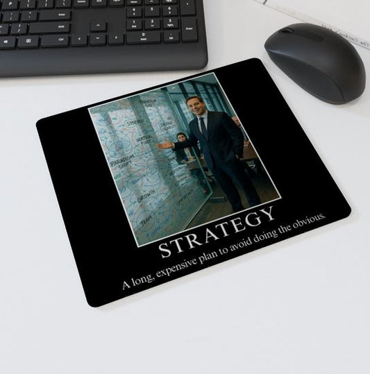 Mouse mat with the text “STRATEGY – A long, expensive plan to avoid doing the obvious” showing a businessman gesturing to a glass wall full of corporate buzzwords and diagrams — parody office desk accessory.